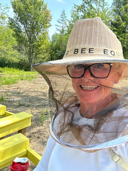 Smiling beekeeper wearing a protective veil beside yellow hive boxes on a sunny day.